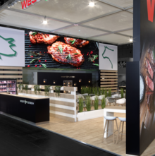 Der Messestand von Westfleisch auf der Anuga Messe. An der Wand hängt ein großes Bild von gegrilltem Fleisch, sowie von stilistischen Zeichnungen eines Schweins uns eines Rinds. Auf dem Gelände stehen viele Stühle und Pflanzen.