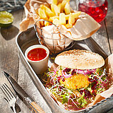 Ein Burgermenü mit Pommes und roter Soße in einem Metallkästchen. Alles steht auf einem Holztisch, auf dem sich Besteck ein Glas mit einem roten Getränk befinden.