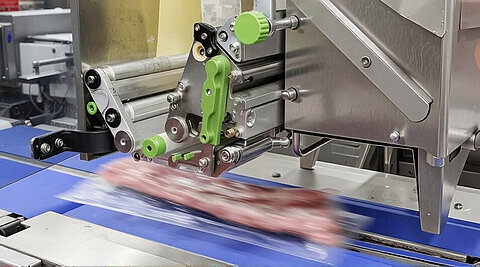 Ein langes Stück Fleisch auf einem Fließband. Ein Verpackungsroboter ist dabei, das Fleisch zu verpacken. 