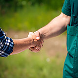 Zwei Personen reichen sich die Hand. Man sieht von ihnen nur die ausgestreckten Arme und einen Teil der Kleidung. Die linke Person trägt ein blau weiß kariertes Hemd. Die rechte Person trägt grüne Landwirtschaftskleidung. 