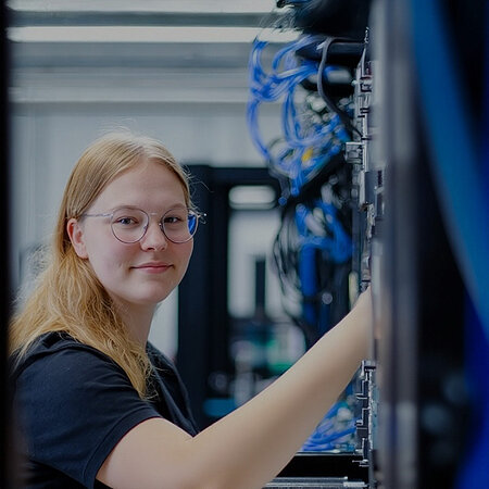 Eine junge, blonde Frau mit Brille arbeitet an den Kabeln eines Servers. 