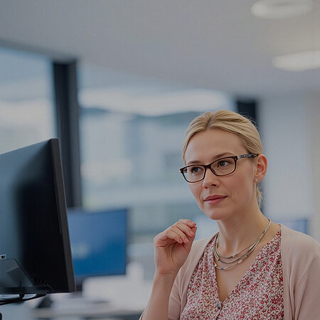 Eine blonde Frau mit Brille in einem rosafarbenen Oberteil sitzt in einem Büro und hat die rechte Hand nachdenklich in Richtung Gesicht erhoben.