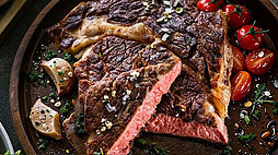 Ein Bild im Querformat von einem gedeckten Tisch von oben fotografiert. Darauf befindet sich ein Teller mit gebratenem Steak, eine Schüssel mit Salat, Besteck und ein bisschen Deko drumherum wie Tomaten und Knoblauch.