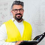 Ein Mann mit Brille und Bart hält eine Mappe in der Hand. Er trägt eine gelbe Warnweste über einem weißen Hemd. Hinter ihm an der Wand sieht man technische Zeichnungen. 