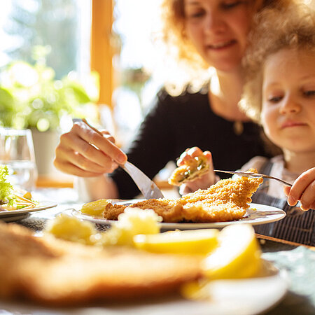 Eine Frau sitzt mit einem Kleinkind auf dem Schoß an einem gedeckten Esstisch. Sie essen ein Schnitzel.