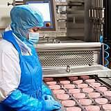 Eine Arbeiterin steht an einem Fließband mit rohen Fleisch Patties. Sie trägt hygienische Schutzkleidung: Kopfhaube, Mundschutz, Schütze und Handschuhe. 