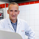 Ein Mann in mittleren Jahren lächelt in die Kamera. Er trägt einen weißen Kittel über einem blauen Hemd. Auf dem Kittel ist das WestfalenLand Logo aufgedruckt. Im Hintergrund sind weiße Fliesen an der Wand. Die oberste Fliese ist jedoch rot. 