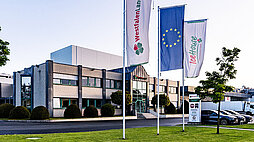 Eine Außenaufnahme der Gebäude von Westfalenland. Vor dem Gebäude wehen die Flaggen von Westfalenland, Icehouse und der EU. Es sind außerdem einige Bäume, Rasen und geparkte Autos auf dem Gelände zu sehen.