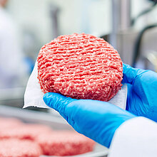Ein roher Burger-Pattie wird von zwei Händen in blauen Lebensmittelhandschuhen gehalten. Im Hintergrund liegen weitere Burger-Patties auf einem Fließband.