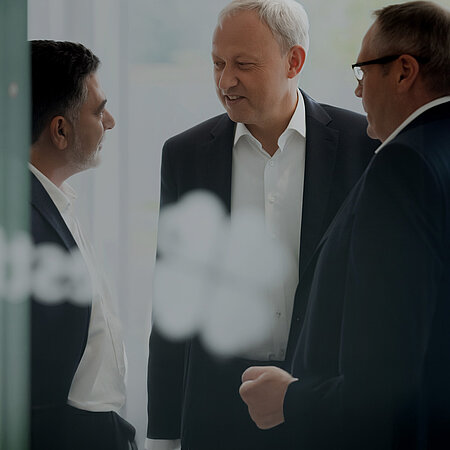 Drei Männer in gehobenem Alter sprechen miteinander. Sie tragen dunkle Anzüge bzw. Business-Kleidung.