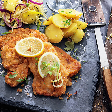 Ein großes Schnitzel auf einer Schieferplatte. Auf dem Schnitzel liegen zwei Schreiben Zitrone. Auf der Schieferplatte liegen neben dem Schnitzel Kartoffeln und Salat.