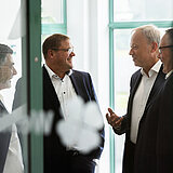 Drei Männer in gehobenem Alter sprechen miteinander. Sie tragen dunkle Anzüge bzw. Business-Kleidung.