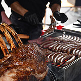Viele Würstchen liegen auf einem großen Grill. Am Grill steht ein Mann in schwarzer Kleidung und mit schwarzen Einweghandschuhe. Man sieht noch ihm nicht den Kopf, nur einen Teil des Oberkörpers und der Beine. Neben dem Grill erkennt man weiteres gebratenes Fleisch, aus dem drei Rippen ragen.