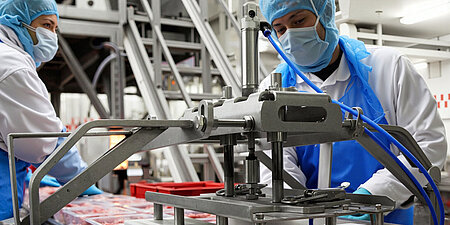 Zwei Arbeiter:innen arbeiten an einem Fließband mit abgepacktem Fleisch. Sie tragen Hygieneschutzkleidung wie Mütze, Maske, Handschuhe und Schürze. 