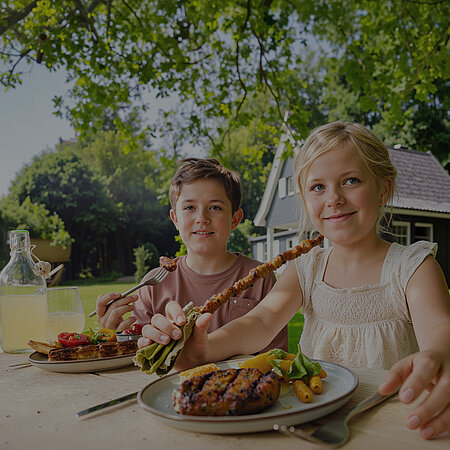 Zwei Kinder, ein Junge und ein Mädchen, sitzen im Freien an einem Tisch und haben einen Teller mit Grillprodukten vor sich. Es befinden sich Grillfleisch und Salat auf den Tellern. Der Junge hält eine Gabel mit einem Stück Fleisch, das Mädchen hält einen Fleischspieß. Im Hintergrund sieht man ein Haus im Grünen. 