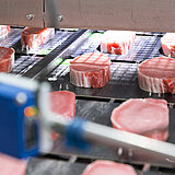 Rohes Fleisch auf einem Fließband, das grade verpackt worden ist. Die Verpackung umschließt das Fleisch eng und Luftdicht. Das Fleisch liegt auf einer Pappe. 