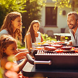 Eine Familie aus Vater, Mutter und zwei Töchtern sitzt im Garten beim Grillen. 