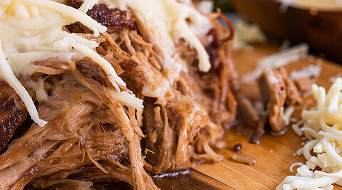 Ein Bild im Querformat mit Pulled Pork mit Käse überbacken in einer Nahaufnahme. Das Fleisch liegt auf einem Holzbrett. Man sieht daneben noch etwas Streukäse, eine Tomate und grüne Blätter von einem Kraut.