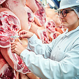 Eine Frau mit einem weißen Kittel und einem weißen Schutzhelm kontrolliert Fleisch in einem Kühlhaus. 