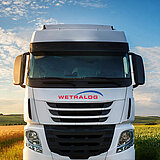 Frontalaufnahme eines Wetralog LKW auf einer Straße. Vorne auf dem Fahrzeug ist das Westfleisch Logo abgebildet. Links und rechts der Straße sind blühende Felder zu sehen. Im Hintergrund ist blauer Himmel.