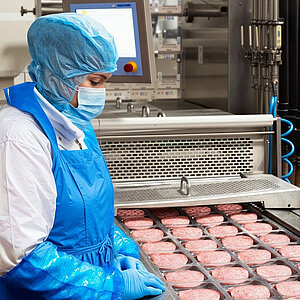 Eine Arbeiterin steht an einem Fließband mit rohen Fleisch Patties. Sie trägt hygienische Schutzkleidung: Kopfhaube, Mundschutz, Schütze und Handschuhe. 