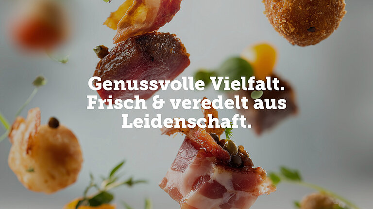 Verschiedene Fleischsorten wie Bacon oder panierte Nuggets fliegen durch das Bild, als würde sie jemand von weiter oben in eine Schüssel werfen. Auch Kräuter wie Basilikum oder gelbe Paprika sind zu sehen und fallen gemeinsam mit dem Fleisch. Im Vordergrund steht in weißer Schrift: Genussvolle Vielfalt. Frisch & veredelt aus Leidenschaft. 