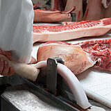 Eine Person schneidet ein großes Stück Fleisch mit einer Maschine. Man sieht von der Person nur einen Teil der weißen Kleidung. 